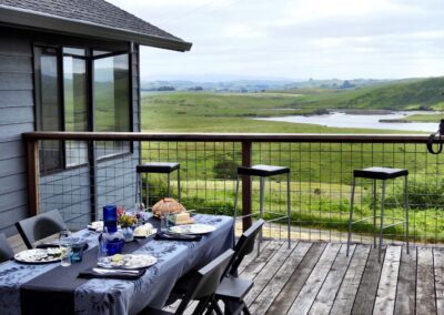 Outdoor dining setup on a wooden balcony overlooking a serene landscape with a river.