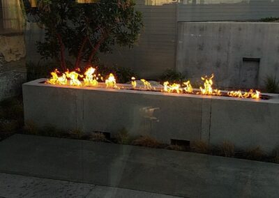 An outdoor modern fire feature with flames along a concrete structure at dusk.