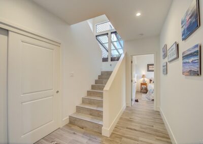 Modern house interior with a staircase and corridor leading to a warmly lit room, featuring light wooden floors and minimalist decor.